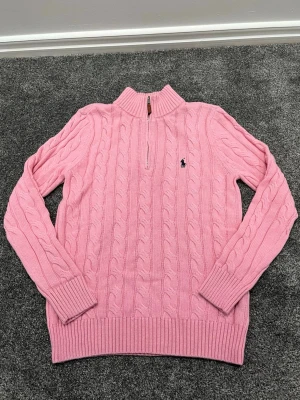 Rosa kabelstickad tröja Polo Ralph Lauren - Kabelstickad rosa tröja från Polo Ralph Lauren med halv dragkedja och hög krage. Tröjan har långa ärmar, ribbade muddar och nederkant samt den klassiska broderade loggan på bröstet. Perfekt för dig som gillar preppy stil och mjuka material.