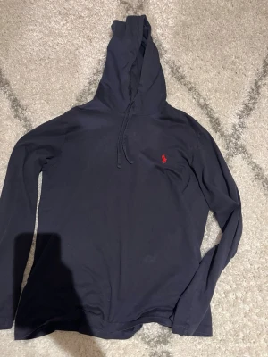 Mörkblå hoodie från Ralph Lauren - Säljer en mörkblå hoodie från Ralph Lauren med den klassiska röda loggan broderad på bröstet. Hoodien har dragsko i huvan och är tillverkad i ett mjukt och bekvämt material. Perfekt för en avslappnad och stilren look.