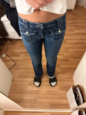 Blå low waist jeans med fickdetaljer - Säljer ett par blå low waist jeans med raka ben och stora fickor framtill med lock och knappdetaljer. Jeansen har klassisk denimlook med kontrastsömmar och bälteshällor. Perfekta för en avslappnad och trendig stil.