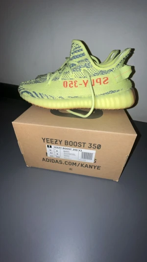 Yeezy Boost 350 V2 Semi Frozen Yellow - Säljer ett par Adidas Yeezy Boost 350 V2 i färgen Semi Frozen Yellow. Skorna har en stickad ovandel i gulgrönt med gråa zebramönster och röd text på sidan. Sulan är i en matchande gul ton och skon har klassiska Yeezy-detaljer och snörning.priset kan diskuteras