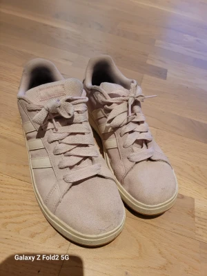 Ljusrosa Adidas sneakers i mocka - Säljer ett par ljusrosa Adidas sneakers i mocka med klassiska vita ränder på sidorna och vit sula. Skorna har lågt skaft och snörning framtill. Adidas-logga i guld på hälen och sidan. Perfekta för dig som gillar stilrena och trendiga sneakers.
