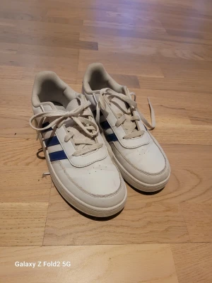 Vita Adidas sneakers med blå detaljer - Säljer ett par klassiska vita Adidas sneakers med blå ränder på sidorna och ljusblå hälpanel. Skorna har lågt skaft, vita snören och ovandel i syntetmaterial. Perfekta för en clean och sportig look.