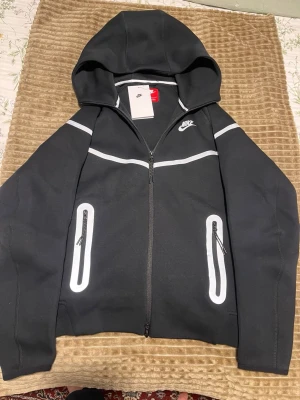 Nike svart loungewear set med huva - Nike loungewear set i svart med vita detaljer. Setet består av en zip-hoodie med huva och två fickor med dragkedja samt matchande byxor med dragsko i midjan och vita ränder längs sidorna. Både tröja och byxor har Nike-logga. Perfekt för chill dagar hemma.