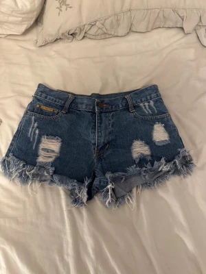 Blå jeansshorts med fransar - Säljer ett par blå jeansshorts från Hollister med slitna detaljer och fransig kant. Klassisk femficksmodell med knapp och dragkedja framtill. Perfekta för sommaren och ger en avslappnad vibe.