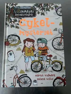 Cykelmysteriet - LasseMajas Detektivbyrå - En färgglad och illustrerad barnbok i serien LasseMajas Detektivbyrå, skriven av Martin Widmark och illustrerad av Helena Willis. Boken har hårda pärmar med tecknade motiv av barn och cyklar på framsidan. Perfekt för unga läsare som gillar mysterier och äventyr.