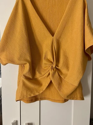 Orange omlottopp med knutdetalj - Trendig orange topp med omlott och knut framtill. Djup v-ringning och korta, vida ärmar ger en avslappnad men snygg look. Tillverkad i ett strukturerat tyg som känns mjukt mot huden. Perfekt för dig som vill sticka ut med färg och form.