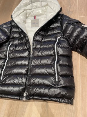Moncler Slim Puffer Svart och Vit  - Snygg svart dunjacka från Moncler med vit insida och huva. Jackan har två stora dragkedjefickor framtill, quiltad design och Moncler-logga på ärmen. Perfekt för kalla dagar och ger en modern, sportig look.