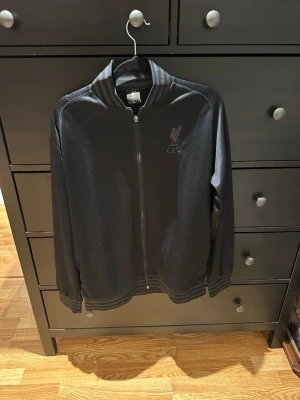 Svart Liverpool FC träningsjacka - Snygg svart träningsjacka med hel dragkedja och ribbade muddar. Jackan har Liverpool FC-logga på bröstet och stor text 'LIVERPOOL F.C.' på ryggen. Tillverkad i ett glansigt, sportigt material som passar perfekt till träning eller som supporterplagg.