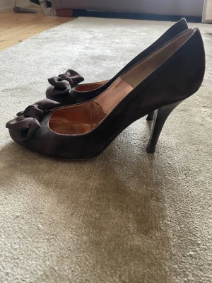 Bruna pumps med rosetter från Marzio - Snygga bruna pumps från Marzio i mocka med rund tå och dekorativa rosetter framtill. Klacken är smal och elegant, vilket ger en klassisk siluett. Perfekta för dig som vill ha en chic och feminin look.