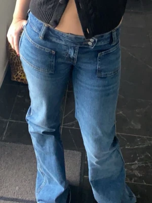 Low waist Blå bootcut jeans med fickor - Snygga blå bootcut jeans med dubbla knappar och stora framfickor. Jeansen har en klassisk denimlook och sitter lågt på höften. Perfekta för dig som gillar en retrostil och vill ha ett par jeans med lite vidare ben.