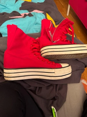 Röda high top sneakers från Yuanbu - Snygga röda high top sneakers från Yuanbu med vita och svarta ränder på sulan. Skorna har dragkedja på sidan, röda skosnören och är tillverkade i canvas. Perfekta för dig som vill sticka ut med färg och cool design.men är mera som en 36 i storlek 