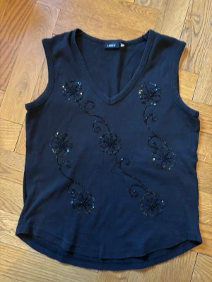 Svart ärmlös topp med blommor - Snygg svart ärmlös topp från Lindex med v-ringning och dekorativa blommor i paljetter och broderi på framsidan. Perfekt till jeans eller kjol för en trendig look. Mjuk och skön bomullskvalitet.