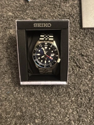 Seiko automatisk klocka svart/silver - Snygg automatisk klocka från Seiko med svart urtavla, silverfärgad länk i metall och detaljer i blått och rött på vridringen. Klockan har datumvisning och tydliga indexmarkeringar. Perfekt accessoar för dig som gillar klassisk men sportig stil. Klockan har lite repor på bandet men inget som stör mig iallafall. Hör av er om ni vill ha fler bilder! 