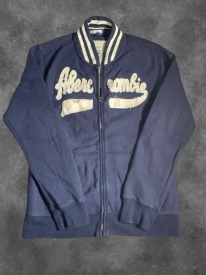 Marinblå tröja från Abercrombie - Snygg marinblå tröja från Abercrombie med dragkedja och vita broderade detaljer på bröstet. Klassisk collegekrage med vita och blå ränder. Tillverkad i mjukt bomullsmaterial och har två fickor framtill. Perfekt för en avslappnad och sportig stil.