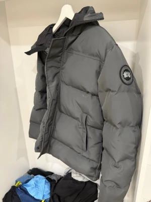 Grå dunjacka från Canada Goose - Snygg grå dunjacka från Canada Goose med svart innerfoder och avtagbar huva. Jackan har två framfickor med dragkedja och en patch med logga på ärmen. Perfekt för kalla dagar och riktigt skön att bära.