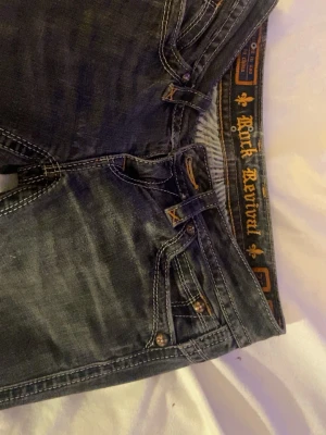 Rock Revival mörk grå jeans nästan typ svarta - Säljer ett par mörkblå jeans från Rock Revival med snygga kontrastsömmar och detaljerade bakfickor med broderade mönster. Jeansen har klassisk femficksdesign, metallknappar och slitna effekter för en cool look. Perfekta för dig som gillar streetstyle.