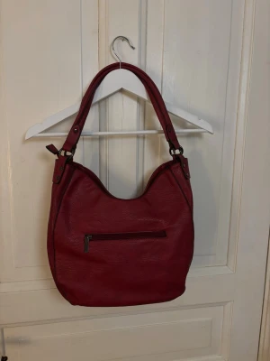 Vinröd handväska i skinnimitation - Snygg vinröd handväska från Eleganci i skinnimitation med en mjuk, rundad form. Väskan har ett ytterfack med dragkedja och justerbara axelband med nitar. Perfekt för dig som vill ha en stilren och rymlig väska med coola detaljer. 