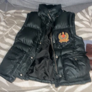 Svart dunväst med broderad logga - Snygg svart dunväst med hög krage och broderad logga med siffran 67 på bröstet. Västen har två stora fickor framtill med knappar och dragkedja. Perfekt för lager-på-lager och streetwear-stil. Den är ganska liten i storleken oxå