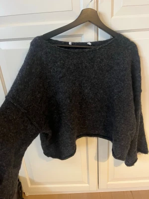 Mörkgrå croppad stickad tröja - Mysig mörkgrå stickad tröja med croppad passform och bred halsringning. Tröjan har långa ärmar och är tillverkad i ett fluffigt, mjukt material som passar perfekt för kyliga dagar.