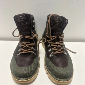 Bruna kängor från Gant med gröna detaljer 42 - Snygga kängor från Gant i brunt läder med gröna gummidetaljer och grov beige sula. Kängorna har metallhål för snörning och bruna snören. Perfekta för dig som vill ha en robust och stilren look.