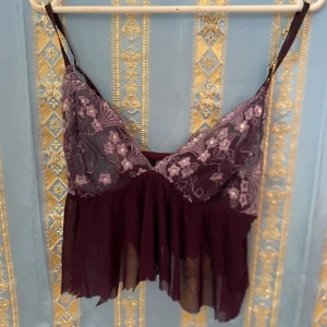 Lila spetslinne med blommor - Säljer ett lila linne med tunna axelband och vacker spets upptill. Toppen har broderade blommor och små paljettdetaljer på bysten samt ett skirt, flowy tyg nedtill. Perfekt för dig som gillar romantisk stil!