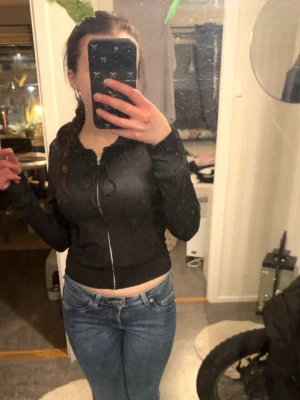 Svart croppad hoodie med dragkedja - Svart croppad hoodie med dragkedja framtill och snörning vid huvan. Långa ärmar och ribbade muddar. Perfekt för en avslappnad och trendig look. Passar till jeans eller leggings. Aldrig använt