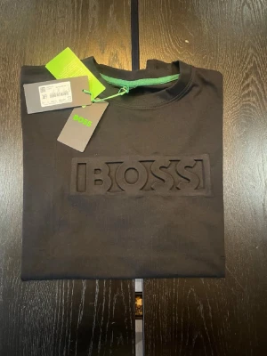 Svart BOSS t-shirt med relieftryck - Snygg svart t-shirt från BOSS med upphöjt relieftryck av loggan på bröstet. Klassisk rund halsringning och korta ärmar. Grön detalj på insidan av kragen och etiketter kvar. Tillverkad i mjuk bomull för skön känsla.
