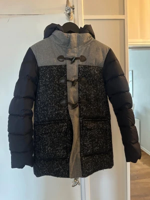 Herno jacka - Stilren dunjacka i blått och grått med huva och snygga träknappar framtill. Jackan har vadderade ärmar, mönstrad kropp och två stora fickor. Perfekt för kalla vinterdagar och ger en modern look. Står ingen storlek på men skulle uppskatta till en S.