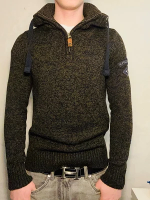 Mörkgrön stickad Superdry tröja - Stickad tröja från Superdry i mörkgrönt melerat garn med hög krage, halv dragkedja och grova blå snören. Tröjan har ribbade muddar och en Superdry-logga på ärmen. Perfekt för dig som gillar en sportig och avslappnad stil.