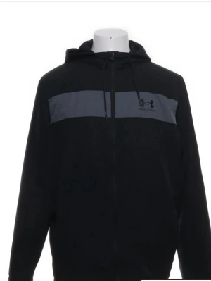 Svart hoodie från Under Armour - Svart hoodie med dragkedja från Under Armour. Tröjan har en mörkgrå panel över bröstet med logga, justerbar huva med snören och långa ärmar. Perfekt för träning eller chill. Tillverkad i mjukt material för extra komfort.