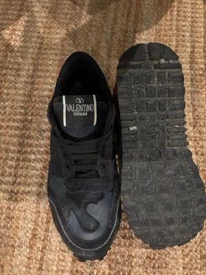 Svarta sneakers från Valentino Garavani - Snygga svarta sneakers från Valentino Garavani med coola kamouflagedetaljer och inslag av mörkrött på hälen. Skorna har en mix av mocka och textil, samt robust gummisula. Perfekta för dig som gillar exklusiva och trendiga sneakers.