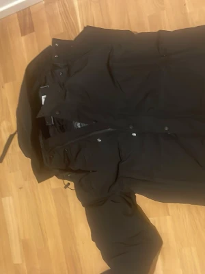 Svart parkasjacka med huva - Säljer en svart parkasjacka med huva och dragkedja samt knappar framtill. Jackan har stora fickor och justerbar dragsko vid huvan. Perfekt för kalla dagar och har en klassisk, stilren look.
