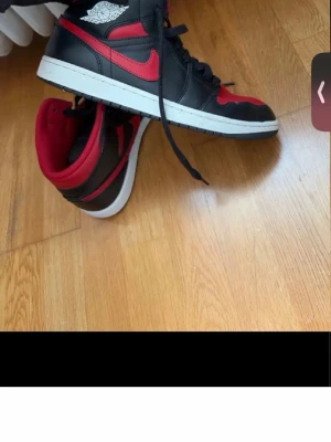 Nike Air Jordan 1 svart/röd sneakers - Säljer ett par klassiska Nike Air Jordan 1 sneakers i svart och rött skinn med vita detaljer och svart snörning. Skorna har den ikoniska Air Jordan-loggan på sidan och en vit platt sula. Perfekta för dig som gillar streetwear och vill sticka ut.