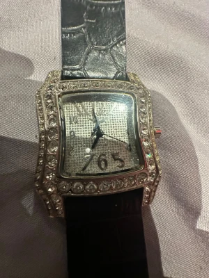 Glittrig fyrkantig klocka med stenar - Snygg klocka med fyrkantig urtavla, glittrande stenar runt boetten och svart läderliknande armband med krokodilstruktur. Urtavlan är silverfärgad och dekorerad med stenar, vilket ger en lyxig känsla. Perfekt accessoar för att sticka ut. Funkar ej!