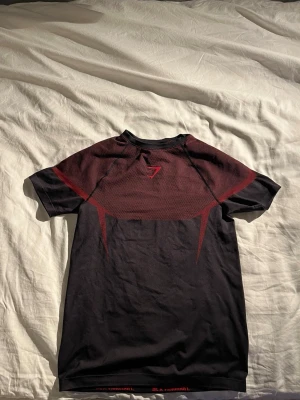 Svart och röd tränings t-shirt Gymshark (onyx v5) - Snygg svart träningströja från Gymshark med röda detaljer och meshpanel över ryggen för extra look. Kortärmad modell i tjockt sportmaterial i bra kvalitet som passar perfekt till gymmet eller löpning. Sportig design med logga på bröstet. Obs!, storleken är Large men passar folk som bär storlek Medium i andra kläder.