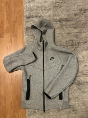 Grå Nike hoodie med dragkedja - Snygg grå hoodie från Nike med hel dragkedja framtill och svarta detaljer. Tröjan har två svarta dragkedjeförsedda fickor och en klassisk Nike-logga på bröstet. Tillverkad i mjukt material och har en stor huva för extra komfort.