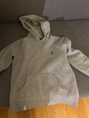 Grå hoodie från Polo Ralph Lauren - Snygg grå hoodie från Polo Ralph Lauren med klassisk huva och magficka. Diskret broderad logga på bröstet. Mjuk och skön bomullsblandning, perfekt för en avslappnad stil. Passar dig som gillar stilrena och tidlösa plagg. Storlek 14-16.