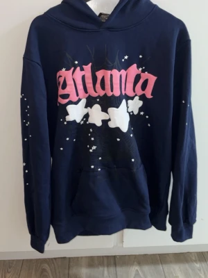 Mörkblå Atlanta hoodie från Sp5der - Mörkblå hoodie från Sp5der med stor rosa 'Atlanta'-text på bröstet och vita stjärnor och fjärilar som detaljer. Klassisk huva och magficka framtill. Materialet är mjuk bomullsmix och designen har en streetwear vibe.