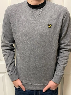 Grå sweatshirt från Lyle & Scott - Säljer en stilren grå sweatshirt från Lyle & Scott i ekologisk bomull. Tröjan har rund hals, ribbade muddar och den klassiska gula loggan på bröstet. Perfekt för en chill och avslappnad look.