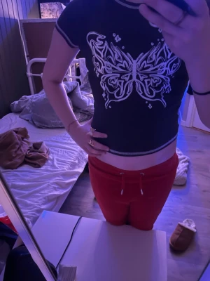 Svart croppad topp med fjärilstryck - Svart croppad topp med vit fjäril på bröstet och kontrastfärgade sömmar. T-shirten har korta ärmar och en tight passform som framhäver midjan. Perfekt för dig som gillar trendiga och unika prints.