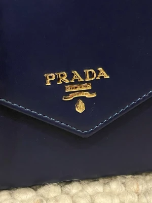 Mörkblå handväska från Prada - Snygg mörkblå handväska från Pr💕💕a i slätt läder med guldfärgade detaljer och kedjerem. Väskan har ett elegant kuvertformat lock med Pra..s logga framtill. Perfekt för dig som vill ha en stilren och lyxig accessoar.