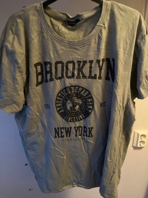 Grå t-shirt Brooklyn från Reaction - Snygg grå t-shirt från Reaction med tryck 'Brooklyn New York' och Athletics Department-märke på bröstet. Klassisk rund halsringning och korta ärmar. Perfekt för en avslappnad streetstyle-look.