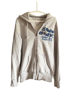 Grå zip-hoodie från Hilfiger Denim - Grå hoodie med dragkedja från Hilfiger Denim. Broderad blå text och siffror på bröstet, två fickor fram och justerbar huva med snören. Mjuk bomullsblandning och ribbade muddar vid ärmslut och nederkant. Perfekt för en avslappnad stil.