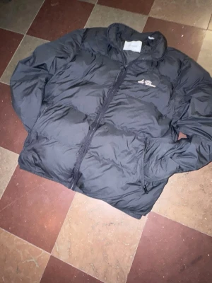 Svart pufferjacka från Les Deux - Säljer en svart pufferjacka från Les Deux med broderad logga på bröstet. Jackan har dragkedja framtill och två sidofickor med dragkedja. Perfekt för kalla dagar tack vare den tjocka vadderingen. Klassisk och stilren design.