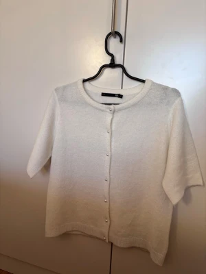 Vit kofta med pärlknappar från 157 - Kortärmad vit kofta från 157 med rund halsringning och pärlknappar framtill. Mjuk och tunn stickning, perfekt för lager-på-lager. Enkel och stilren design som passar till många outfits. Inga frl på den. 