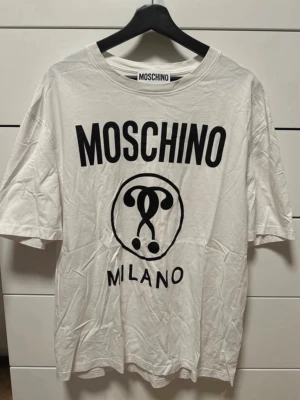 Vit Moschino Milano t-shirt - Vit t-shirt från Moschino med stor svart logotyp och Milano-tryck framtill. Klassisk rund halsringning och korta ärmar. Perfekt för dig som gillar statement-plagg och vill sticka ut med en ikonisk design.
