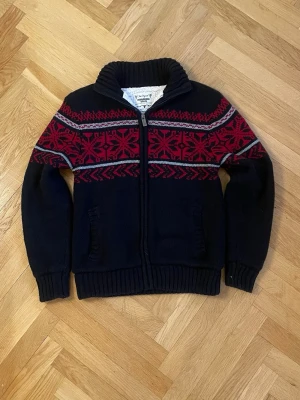 Svart kofta med rött mönster Cedarwood - Mysig svart stickad kofta från Cedarwood State med dragkedja och rött nordiskt mönster över bröst och armar. Koftan har hög krage, två fickor framtill och ribbstickade muddar. Perfekt för kalla dagar och snygg till jeans. Riktigt trendiga nu också! Hör av dig om mer frågor!🙌