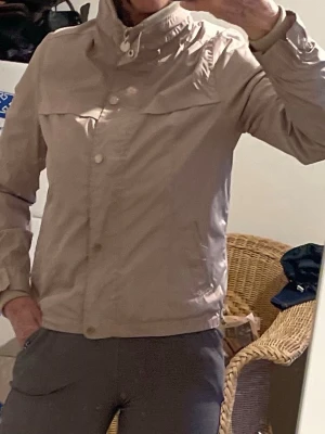 Beige vindjacka med hög krage - Stilren beige vindjacka med hög krage och knäppning framtill. Jackan har långa ärmar, diskreta fickor och är tillverkad i ett lätt, vindtätt material. Perfekt för blåsiga dagar och enkel att matcha med olika outfits.storlek som smal eller medium (personen på bild har medium)