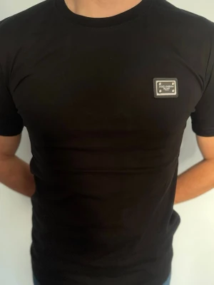 Svart Dolce & Gabbana t-shirt - Snygg svart t-shirt från Dolce & Gabbana med klassisk rund hals och en liten metallbricka med logga på bröstet. T-shirten har korta ärmar och är gjord i mjuk bomull för en bekväm passform. Perfekt till jeans eller shorts.