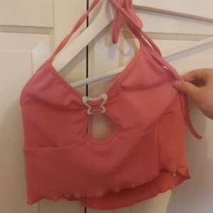 Rosa croppad linnetopp med fjäril - Supersöt rosa croppad linnetopp med ribbad struktur och knyt i nacken. Framtill finns en vit marmorerad fjärilsdetalj och en rund cut-out. Toppen har vågig kant nedtill och är ärmlös, perfekt för att sticka ut med en unik look.